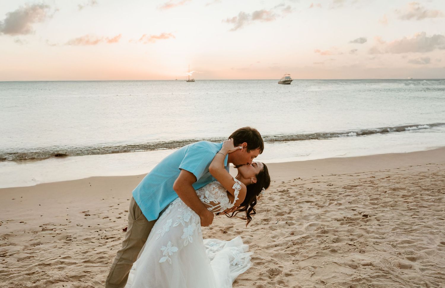 Couple sharing a romantic dip kiss on a sandy beach in St. Lucia at sunset with ocean waves and boats in the background.