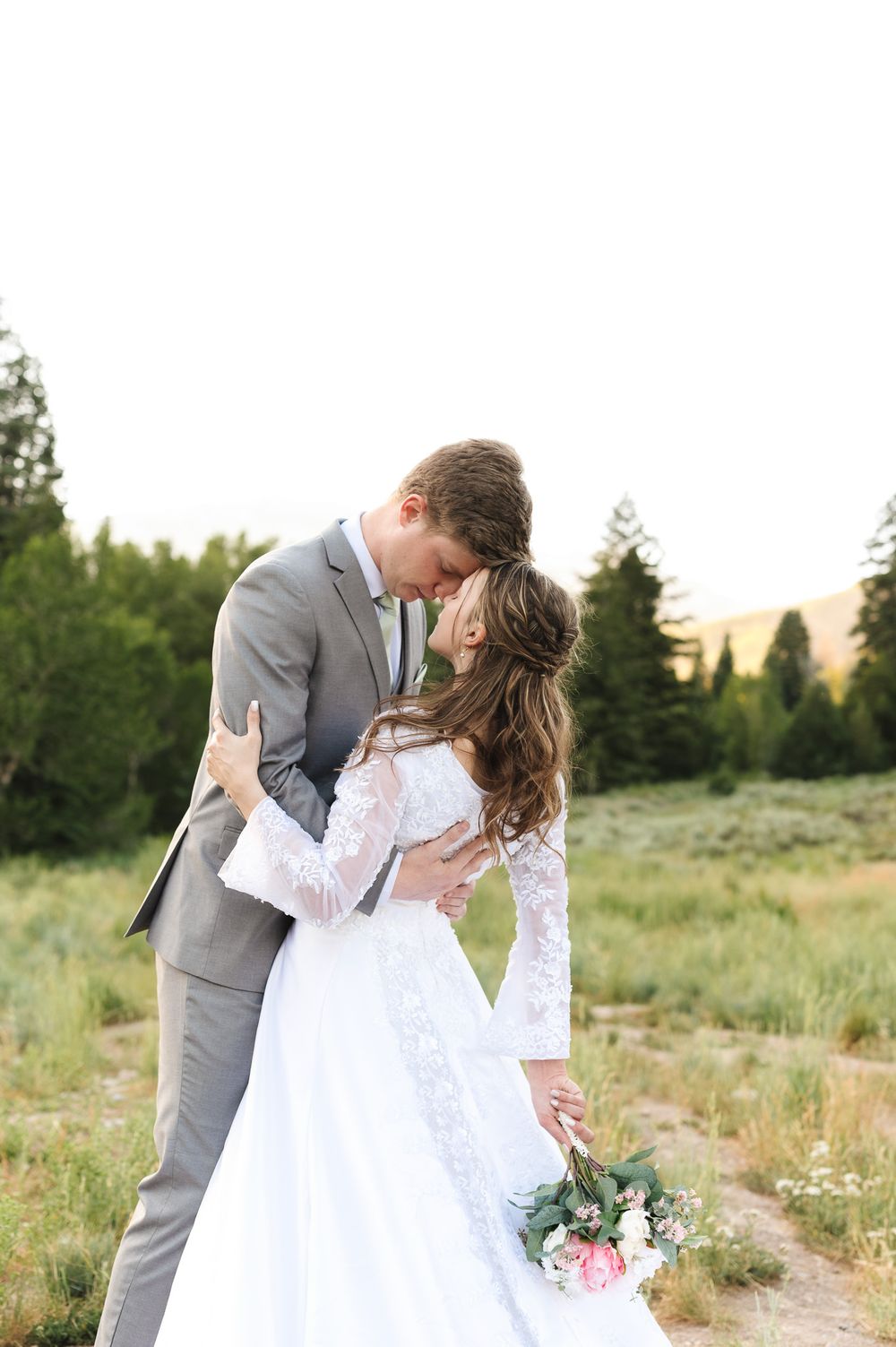 A romantic wedding couple embraces in a scenic meadow with wildflowers and mountains during golden hour.