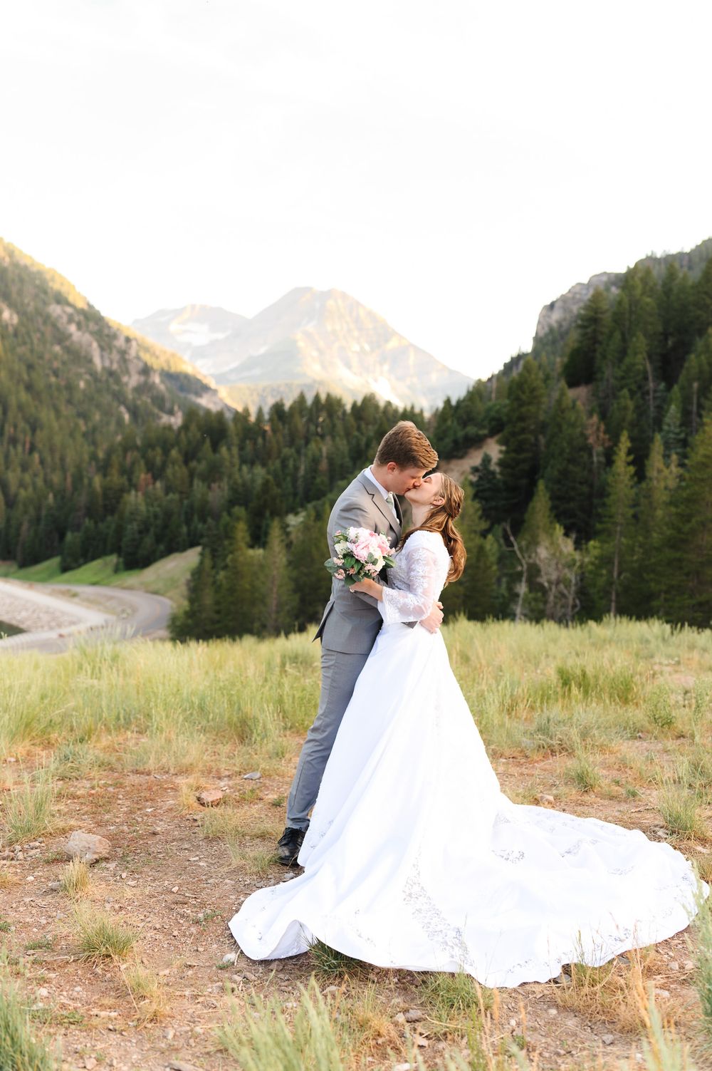 A romantic wedding couple embraces on a mountainside with evergreen forests and peaks in the scenic background.
