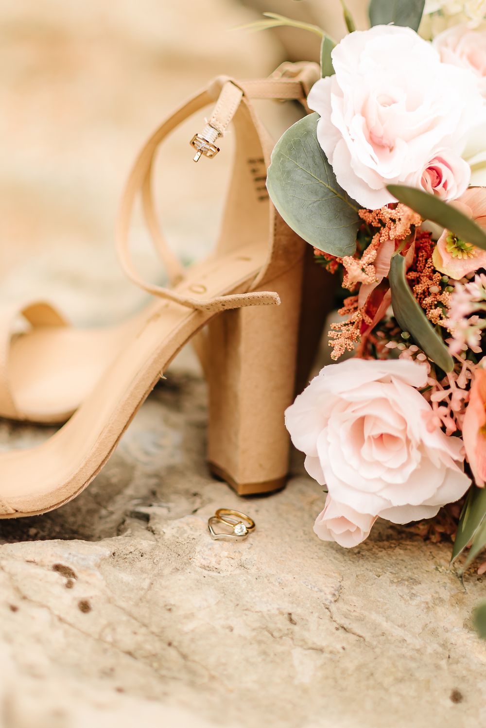 Wedding details featuring blush pink roses, beige heels, and gold wedding rings on a textured stone surface.