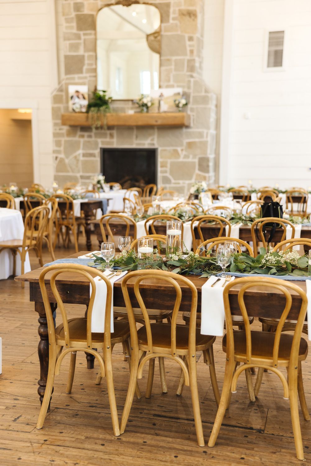 Elegant wedding reception tables set with wooden chairs, white linens, and greenery centerpieces in a rustic stone venue.