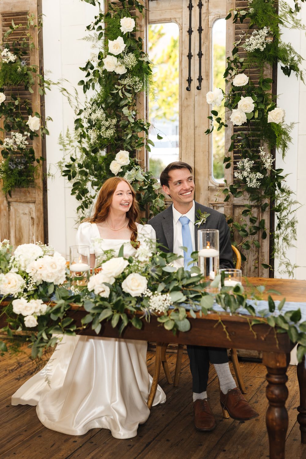 Wedding reception seating area decorated with climbing white roses and greenery along wooden beams.