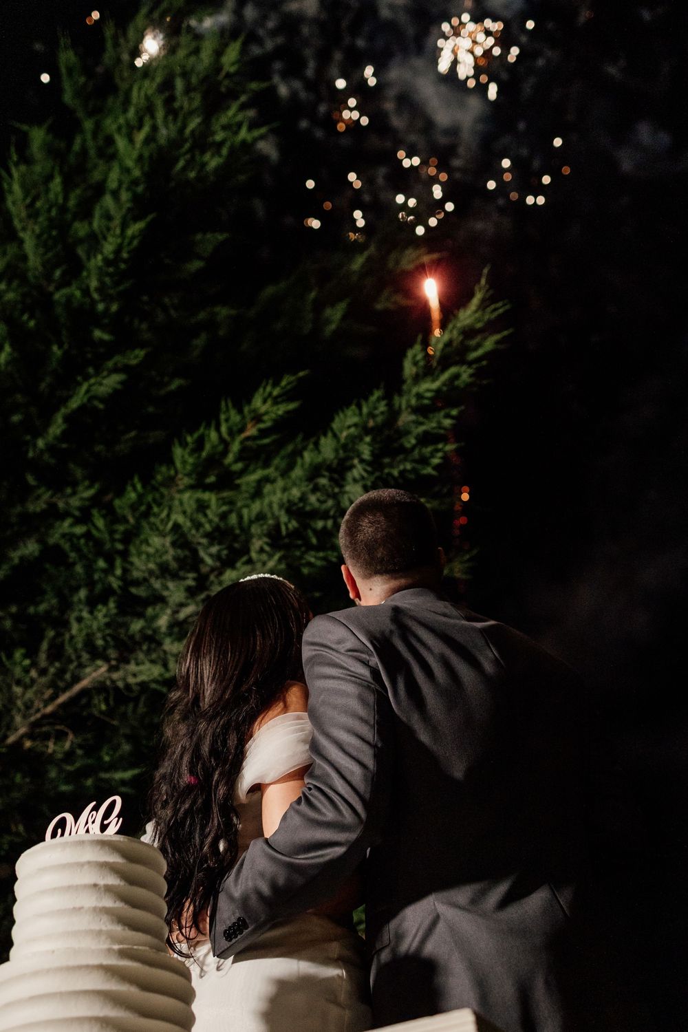 A couple stands close together at night surrounded by twinkling lights and evergreen trees in a romantic setting.