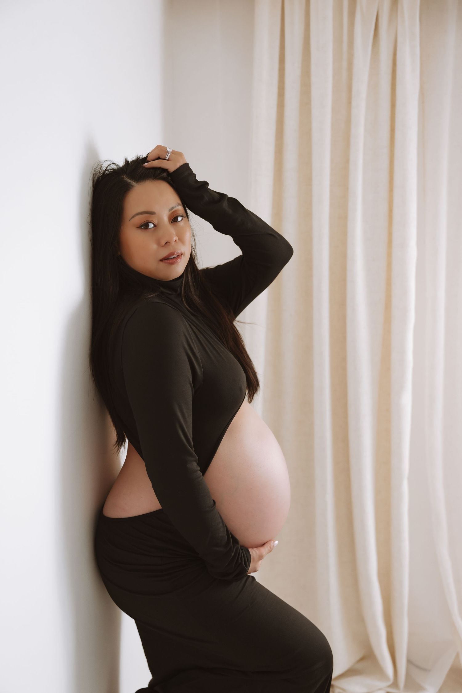 A pregnant woman in a black long sleeve top poses against a white curtain background in a photo series.