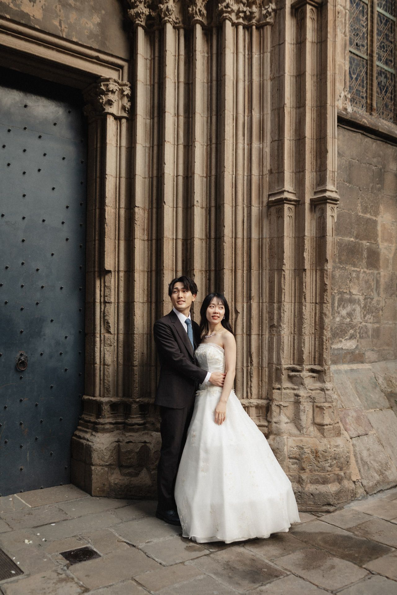 A couple in wedding attire poses in front of ornate Gothic stone columns and a blue door of a historic cathedral.