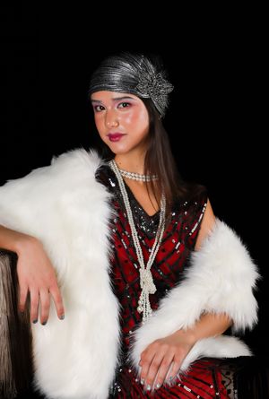 Girl in a red flapper dress, a white shall, a pearl necklace and a silver turban, smiling and looking at the camera