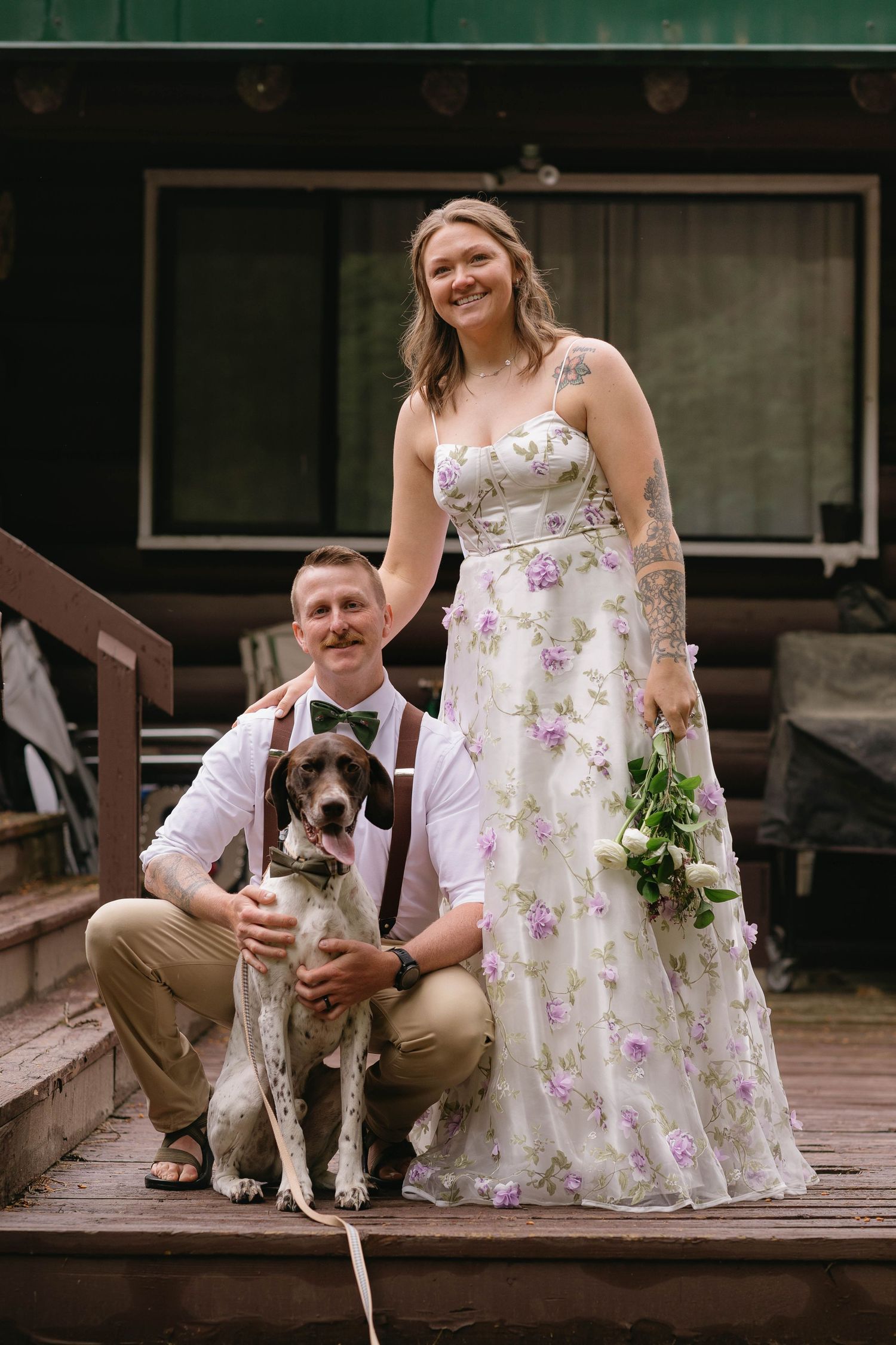 A couple poses with their dog on wooden steps outside a rustic green cabin wearing wedding attire.