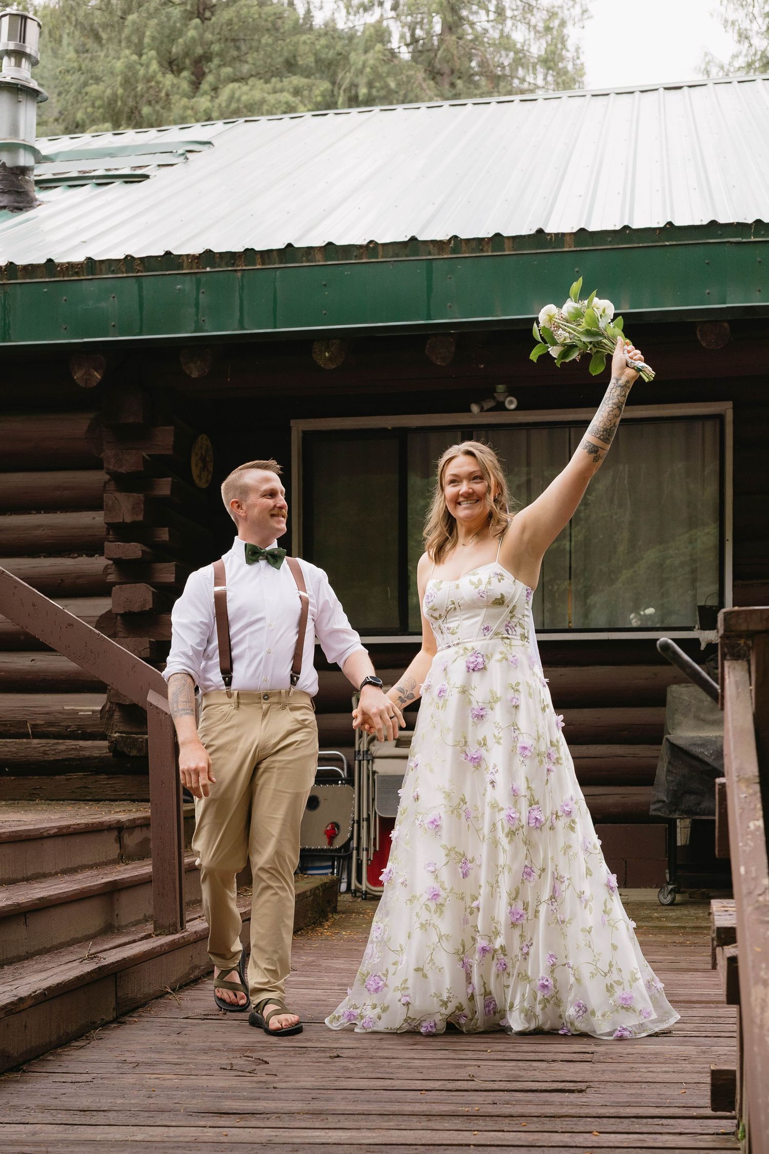 A couple celebrates on a rustic wooden deck outside a log cabin, with the bride in a floral dress raising her bouquet.