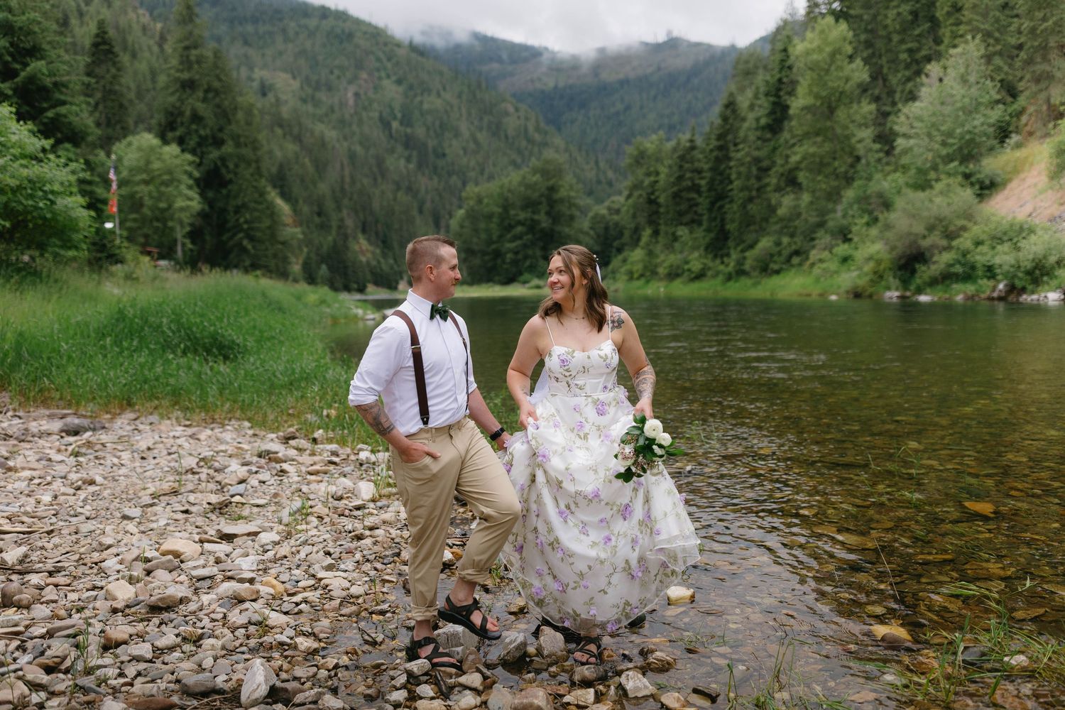 Newlyweds enjoy a romantic moment by a scenic mountain lake surrounded by evergreen forest and rocky shores.