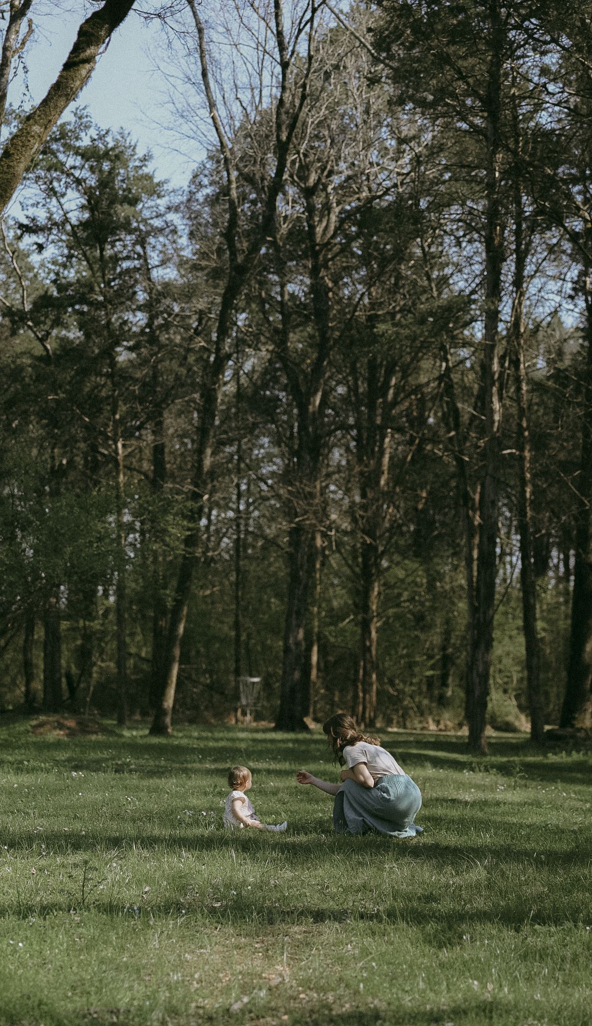 Mother crouching down to interact with her toddler on a grassy woodland clearing.