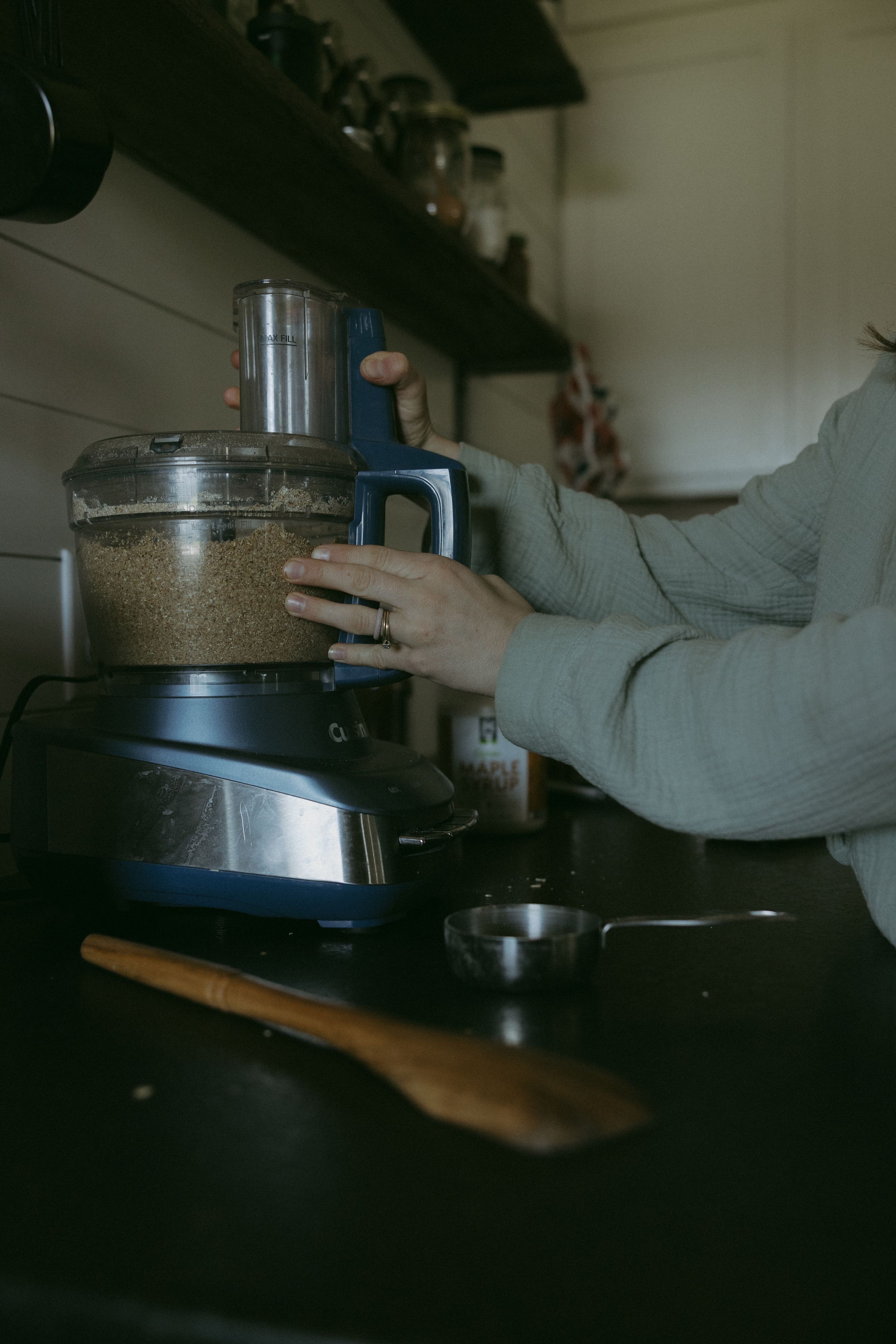 Woman using a food processor in a rustic kitchen with wooden spoon and strainer on dark countertop.