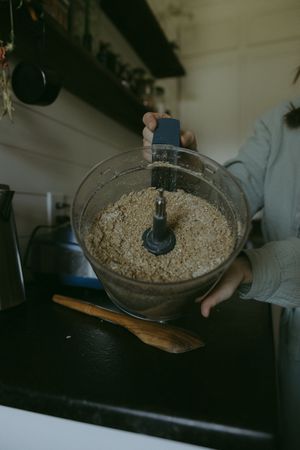 Hands holding food processor bowl with finely ground flour or meal in a dark kitchen setting.