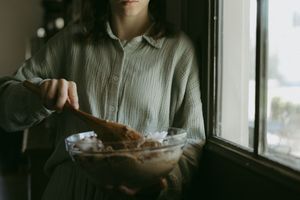 Woman in sage green linen shirt mixing ingredients in glass bowl with wooden spoon by window.