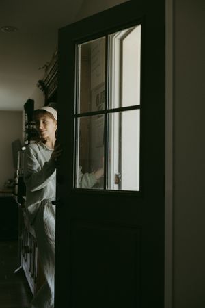Young woman in white cap and apron opening a dark door with glass panels in dim interior.