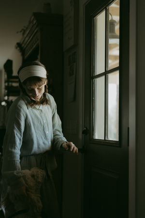 Young girl in headband and blue sweater opening a dark interior door in a dimly lit room.