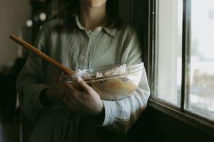Woman in sage linen shirt holding glass mixing bowl with wooden spoon near bright window.