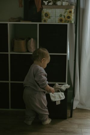 Toddler in lavender outfit pulling cloth from a dark cubby shelf unit in a dim room.