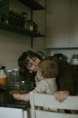 A mother tenderly kisses her baby sitting in a high chair at a dark kitchen counter.