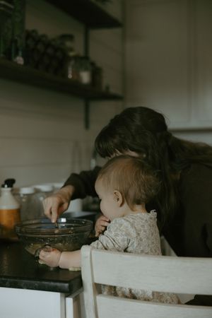 A mother and baby explore a mixing bowl together at a dark kitchen counter.