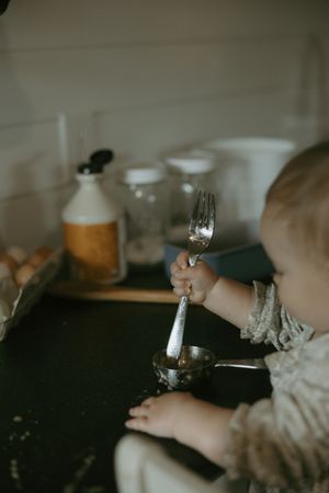 A baby holds a fork and spoon on a dark kitchen counter surrounded by baking ingredients.