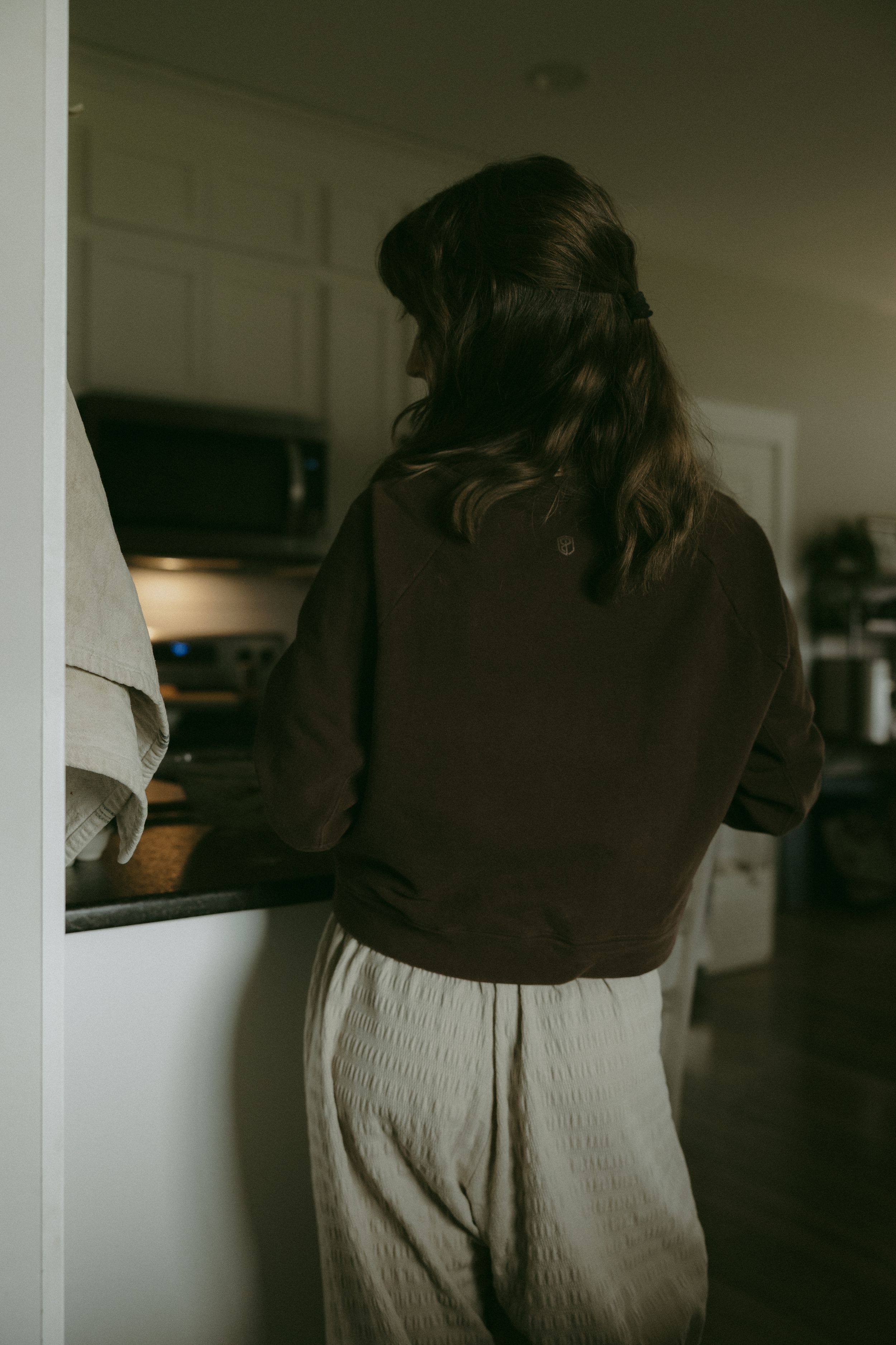 A woman in a brown jacket and cream pants stands at a kitchen counter from behind.
