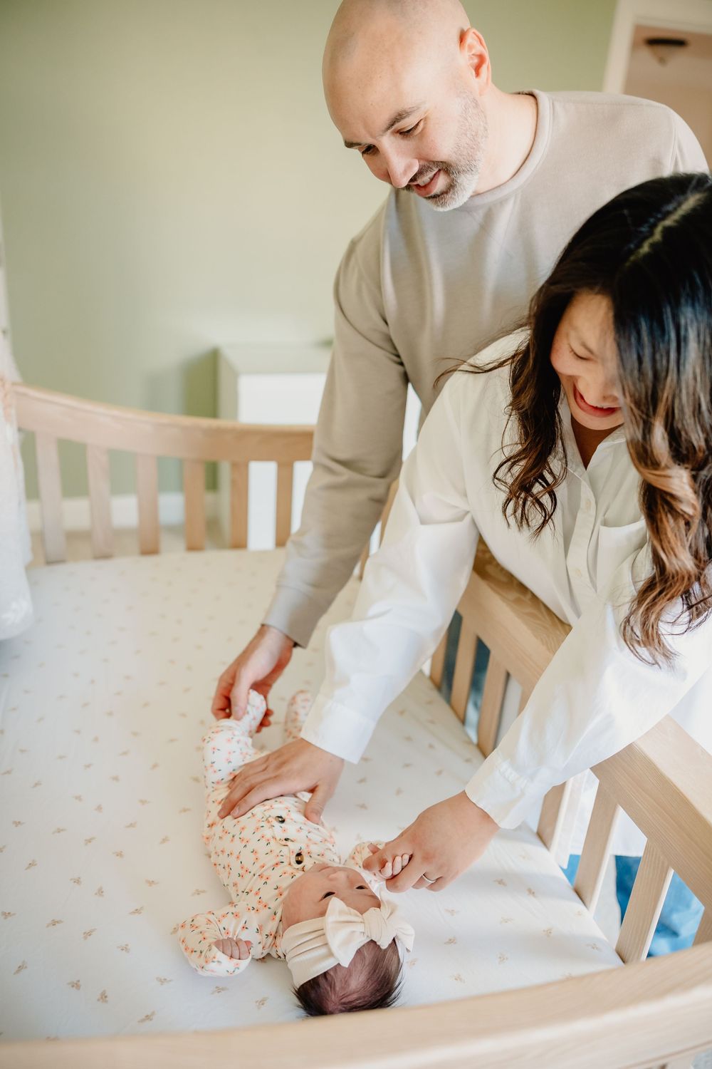 Parents smiling over newborn baby lying in wooden crib, dressed in floral onesie with bow headband.