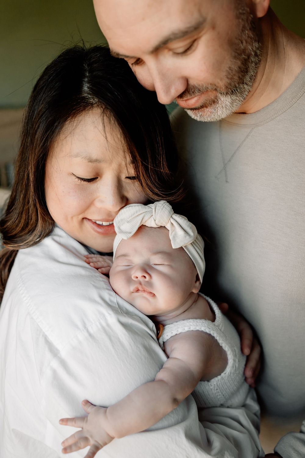Parents tenderly hold their sleeping newborn baby girl wearing a white bow headband and knit outfit.