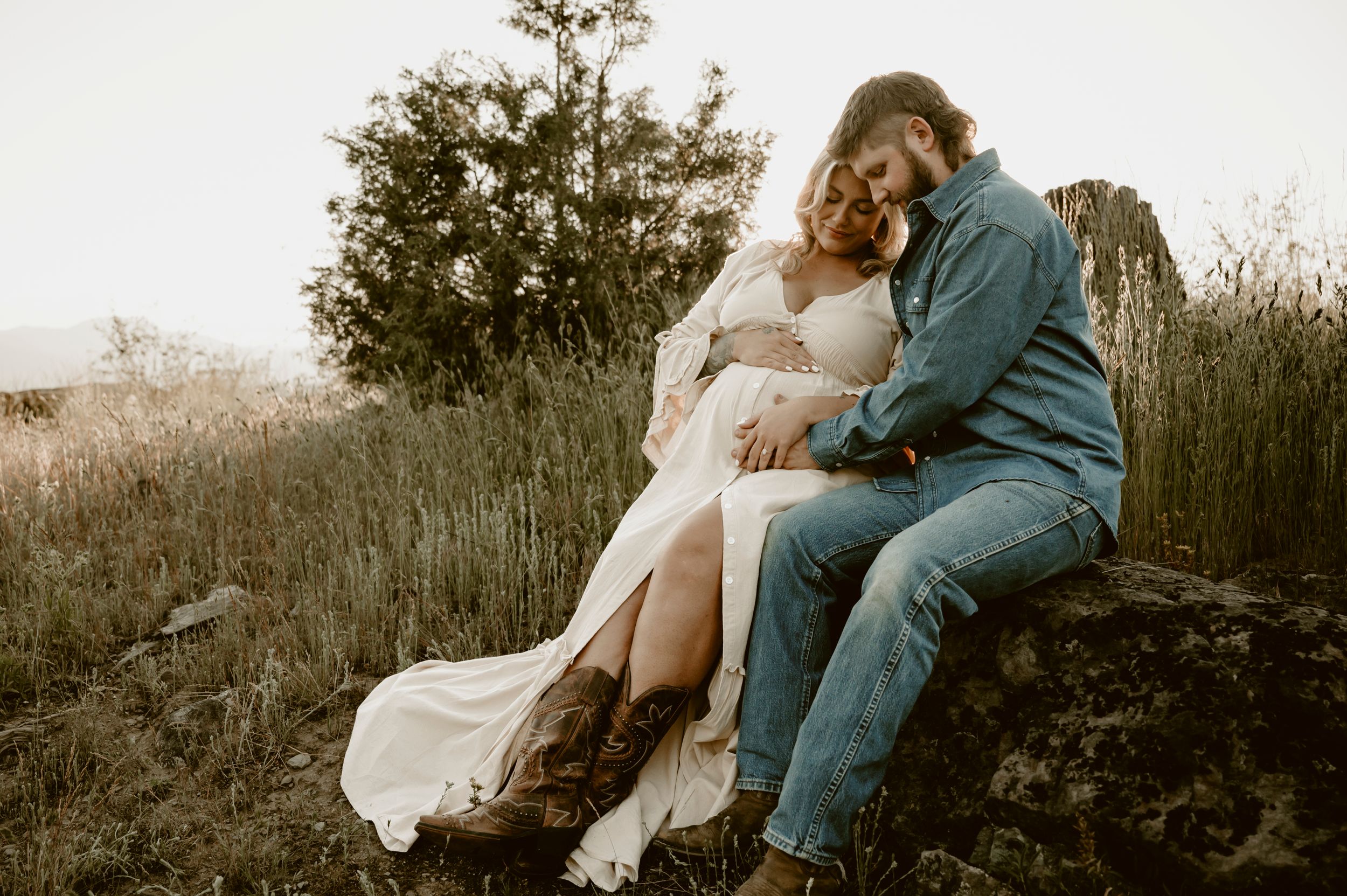 Couple sharing a tender moment on a rock in a field, woman in white dress with baby bump, man in denim jacket.