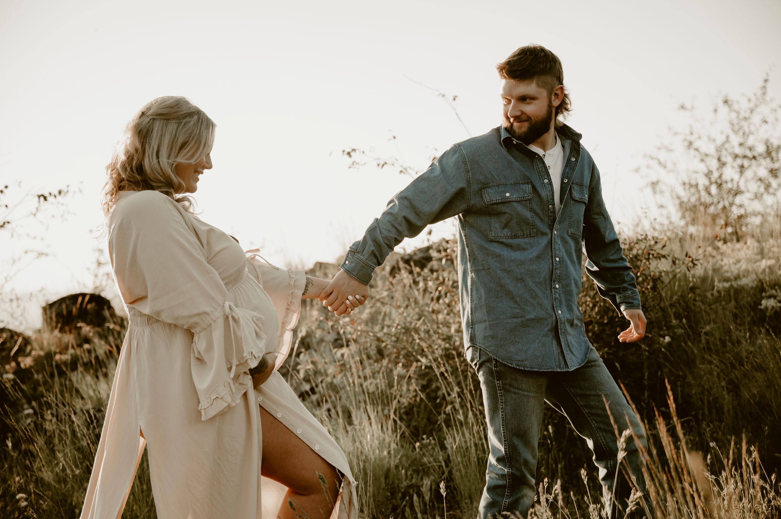 A bearded man in a denim shirt helps a blonde woman through tall grass during a golden hour outdoor photoshoot.