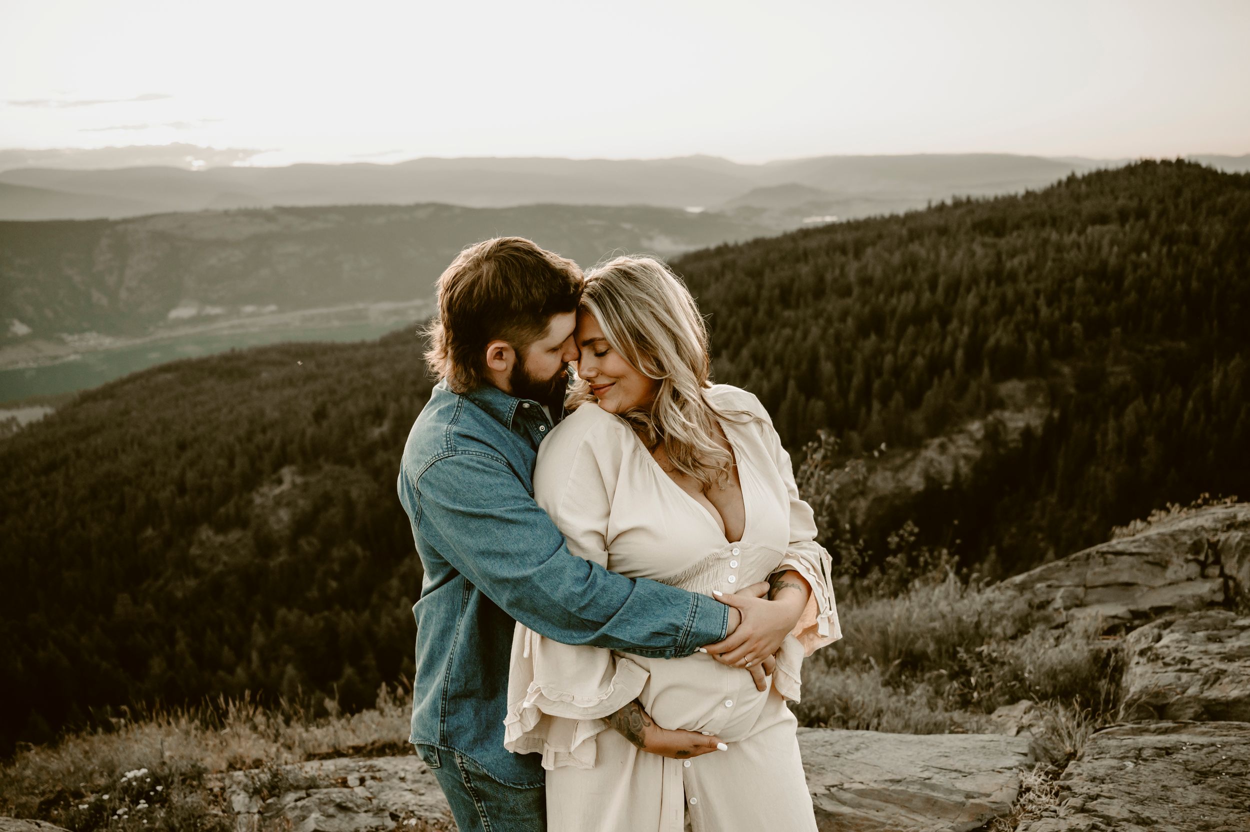 Couple embracing on mountain overlook at sunset, man in denim holding pregnant woman in white dress.