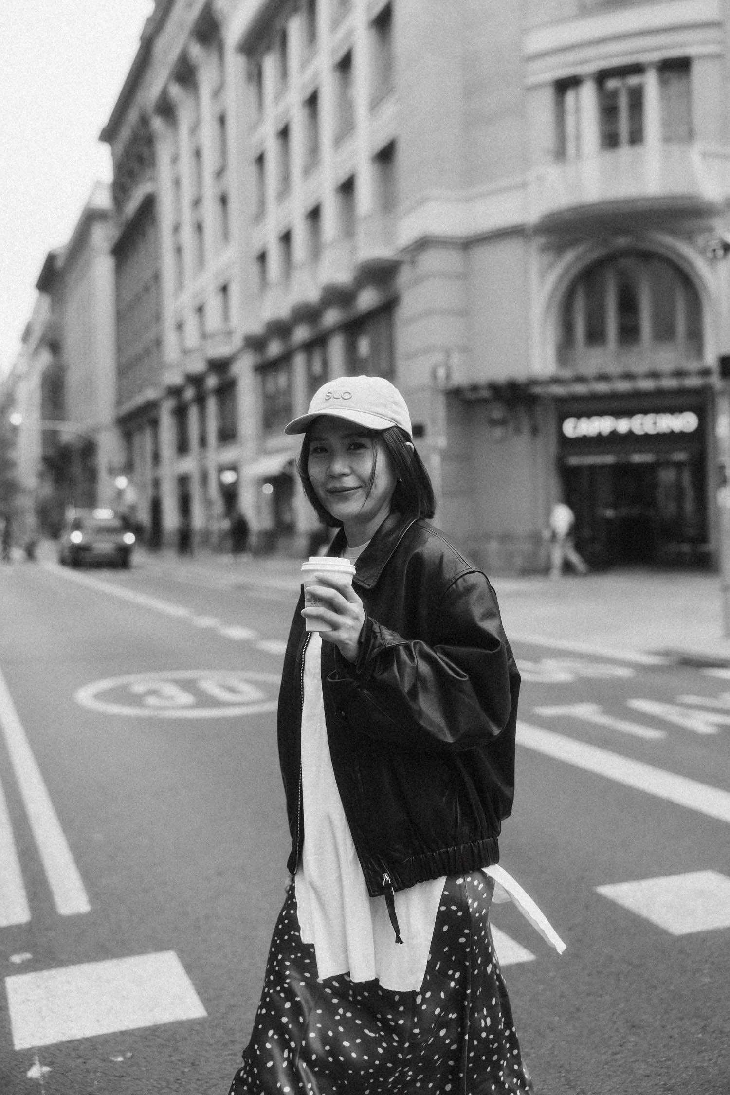 Black and white photo of a smiling woman in a cap and jacket holding a coffee cup while crossing a city street.