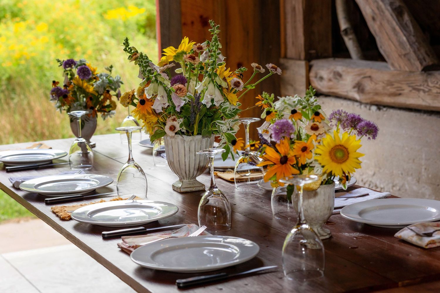 Rustic outdoor dining table set with wildflower bouquets featuring sunflowers, plates, and glassware for an elegant garden event.
