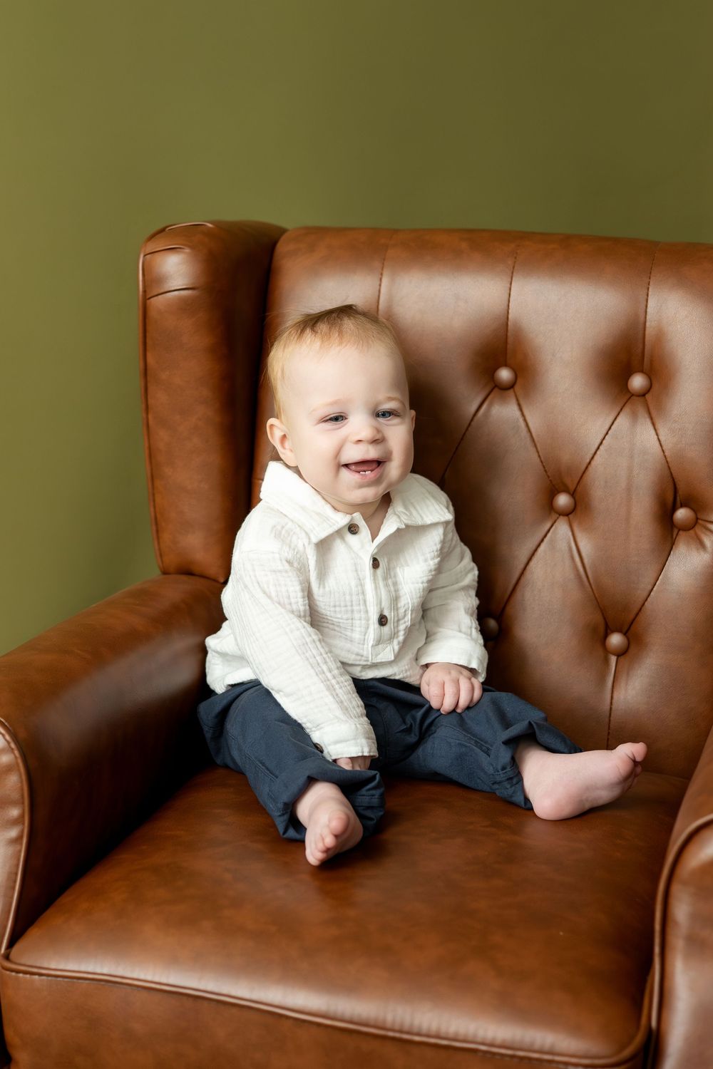 Happy baby in white shirt sits smiling in a brown leather tufted armchair against green wall.