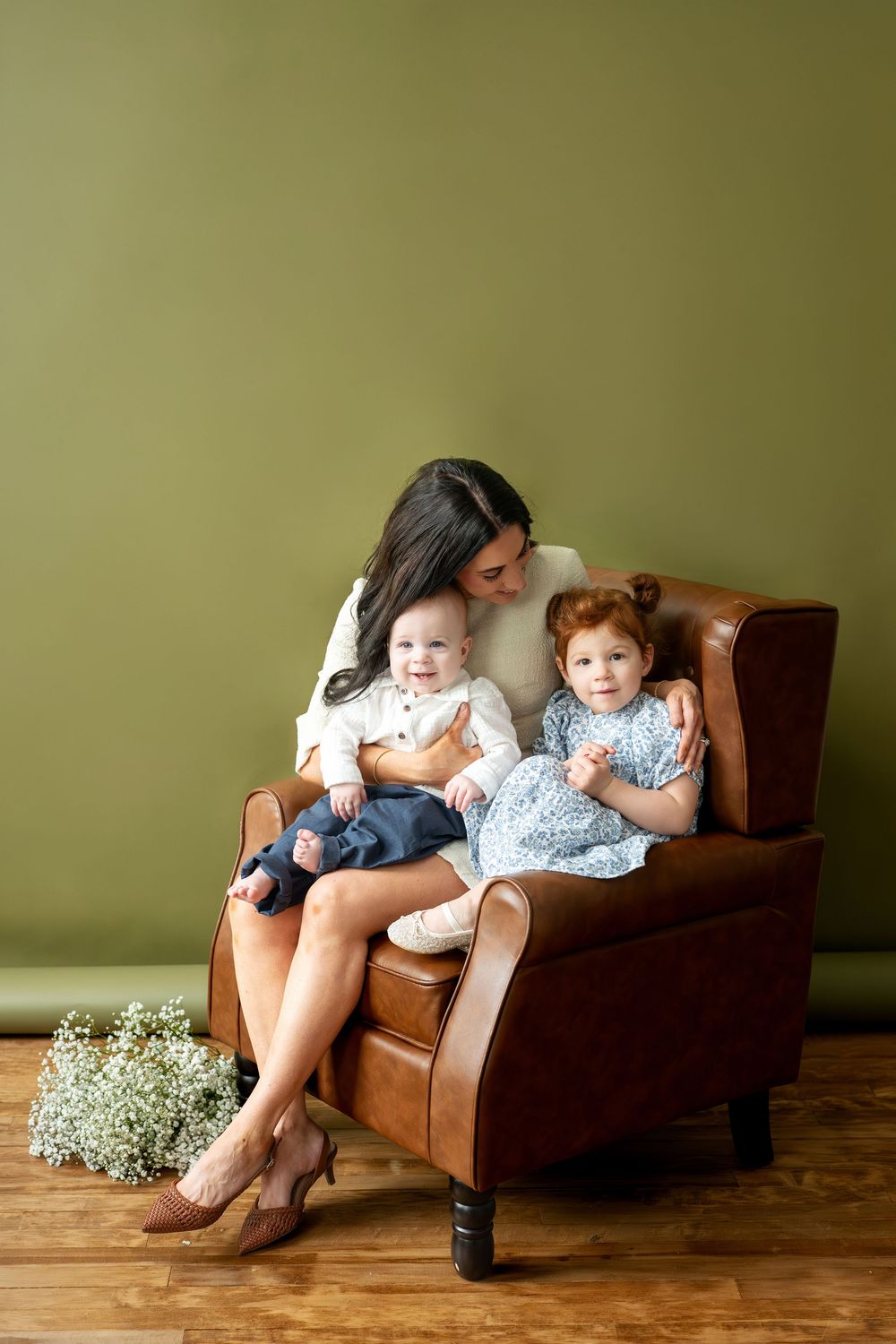 Mother and two young children sitting together in brown leather armchair against olive green wall.