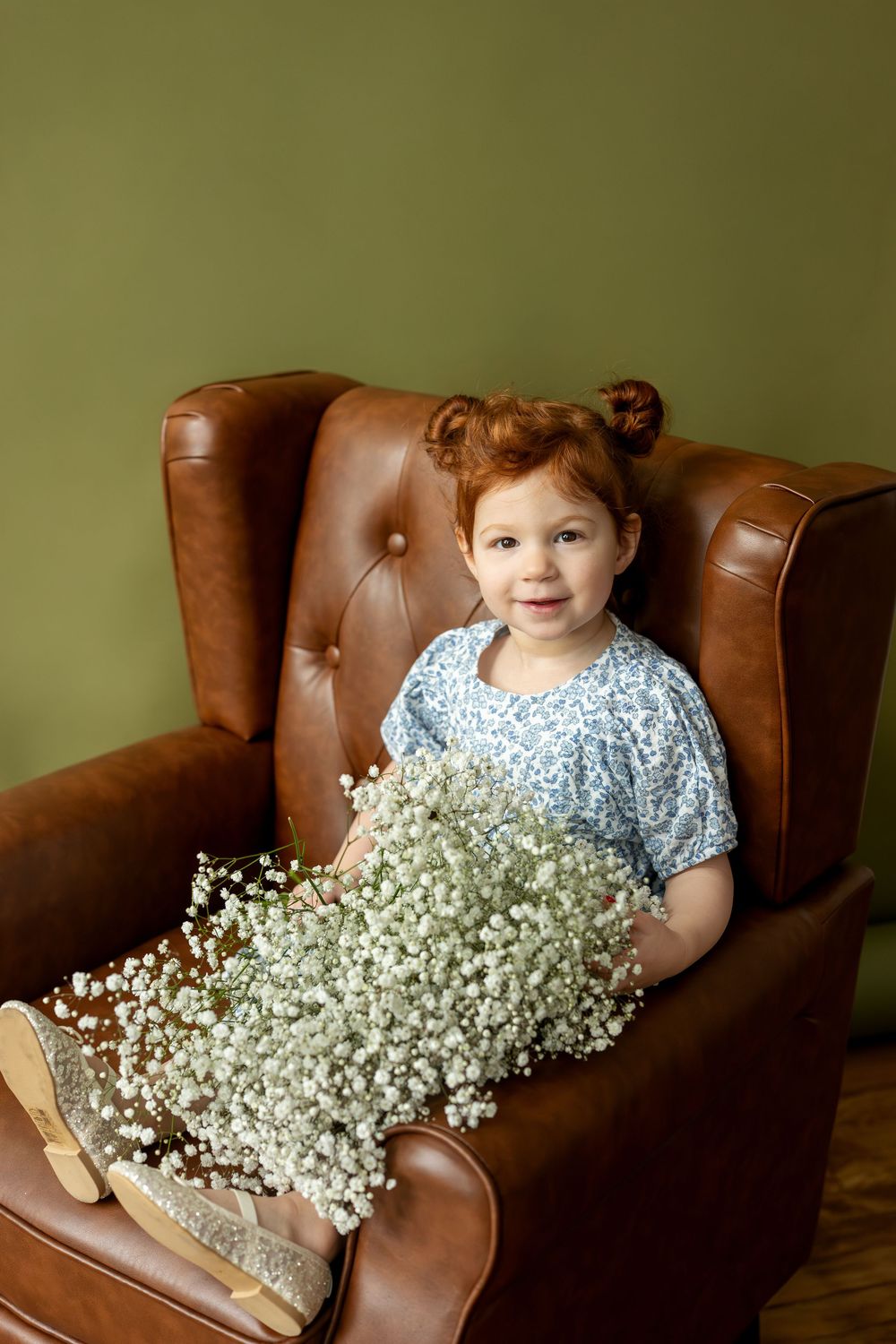 Cute redhead toddler girl holding large bouquet of white baby's breath flowers in brown leather chair.
