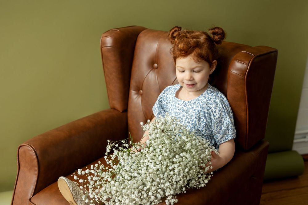 Toddler girl with red hair sitting in brown leather chair holding large bouquet of white baby's breath flowers.