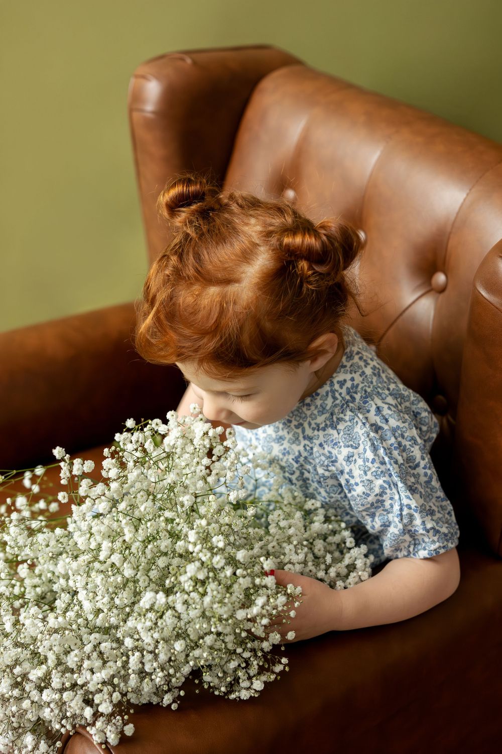 Cute redhead toddler girl smelling white baby's breath flowers while sitting in a brown leather armchair.