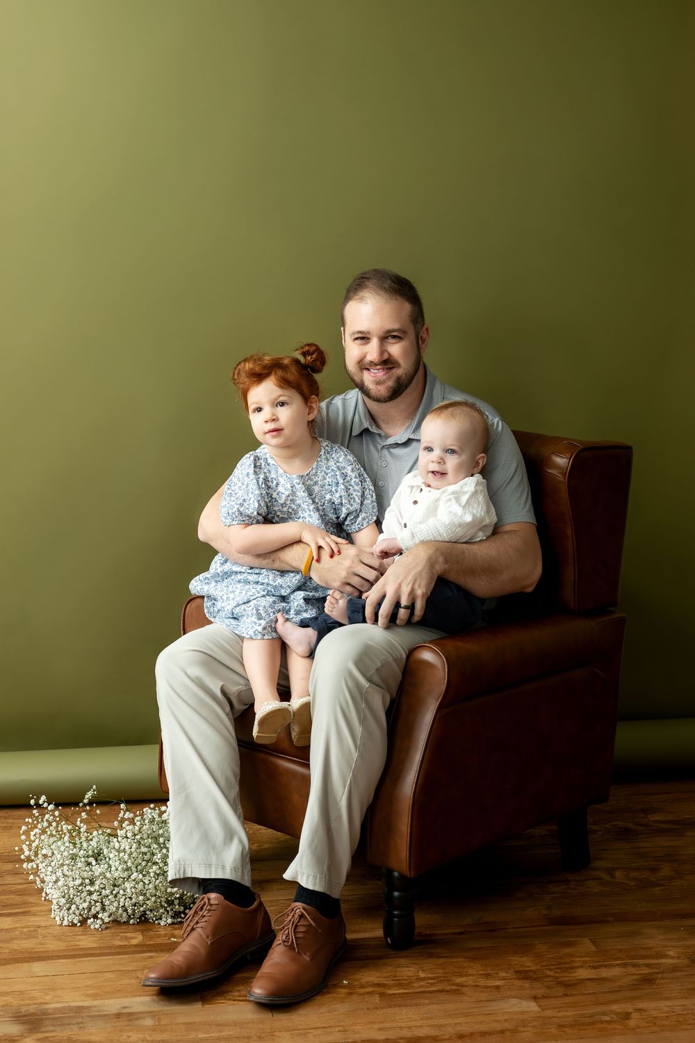 Smiling father sitting in leather armchair holding redhead toddler daughter and baby boy against green wall.