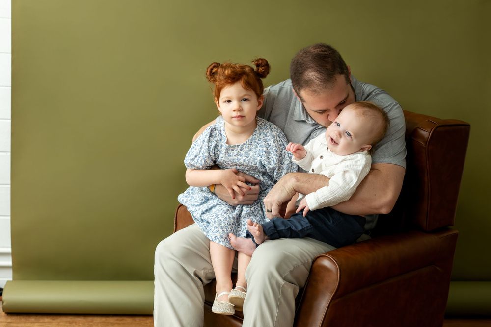 Father sitting in brown leather chair kissing baby while redhead toddler daughter sits beside them.