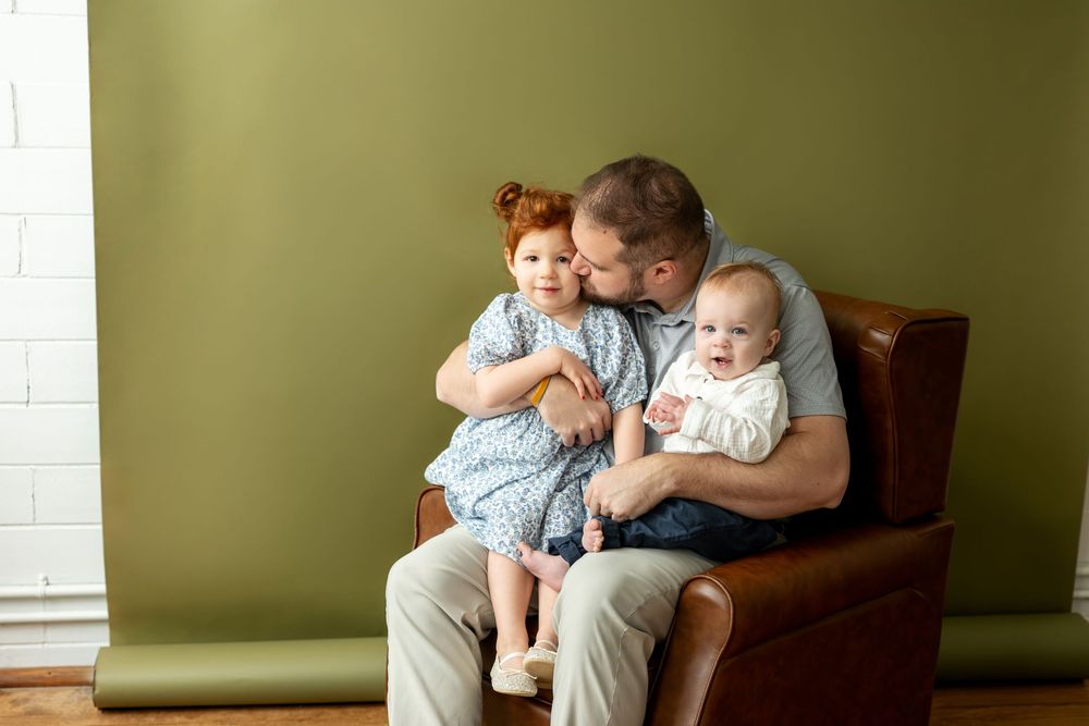 Dad kissing young redhead daughter while holding baby in brown leather armchair against green wall.