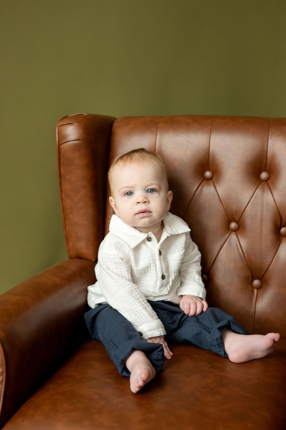 Baby boy in white shirt and navy pants sitting on brown tufted leather chair against olive green wall.