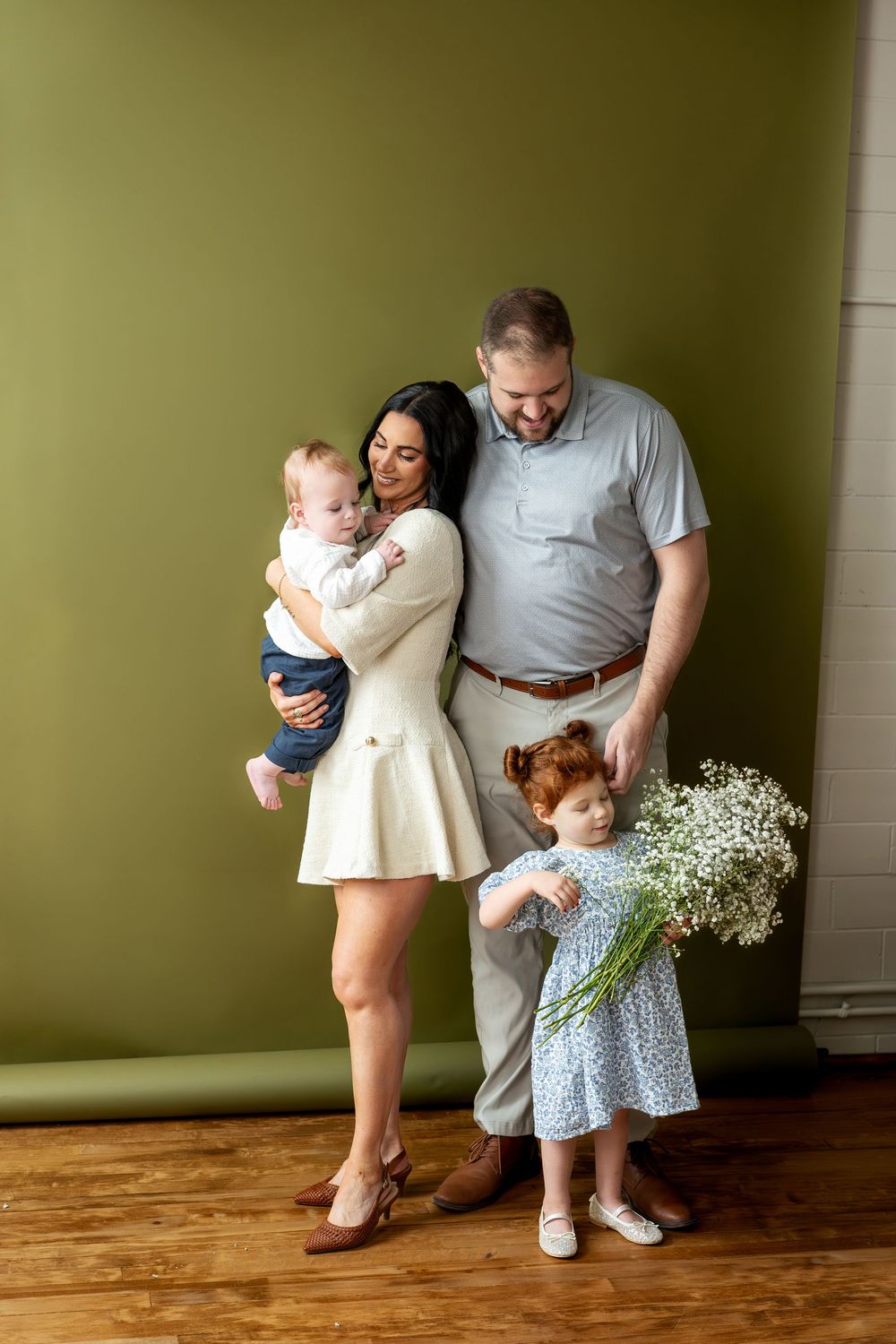 Happy family with brunette mom holding baby and dad standing with redhead daughter holding white flowers.