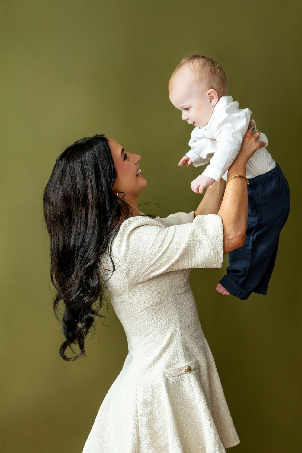 Brunette mother in white dress lifting smiling baby boy up in the air against olive green background.