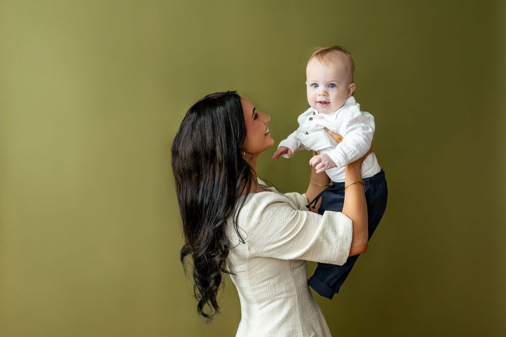 Mother holding baby in white outfit, smiling against olive green background.