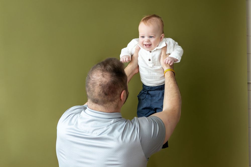 Father lifting smiling baby in white outfit against olive green background.