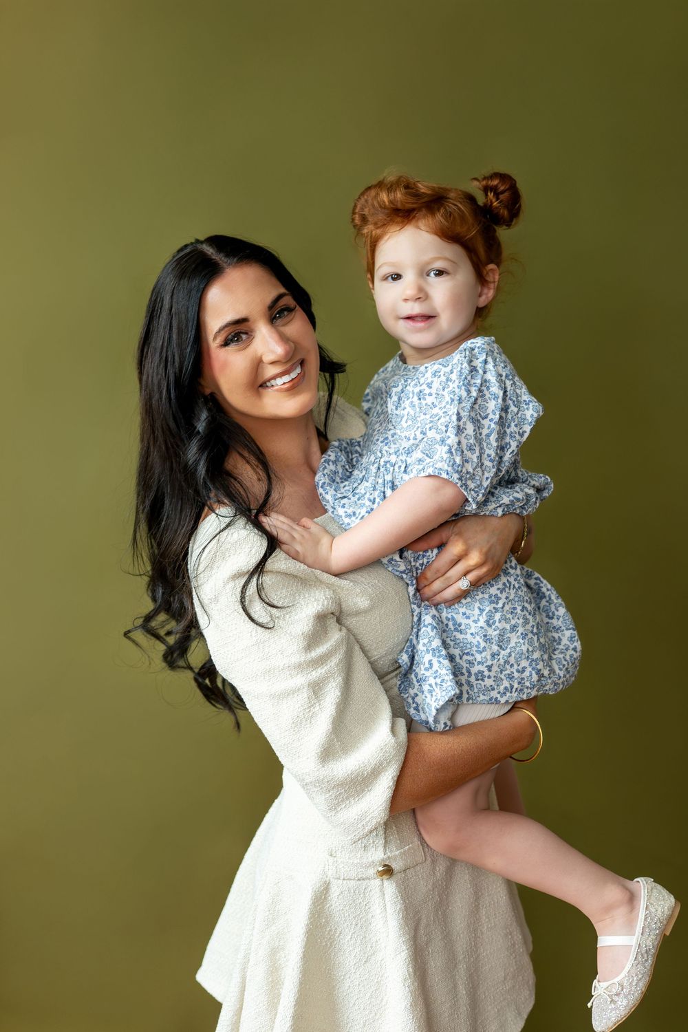 Smiling brunette mother holding redhead toddler girl in blue floral dress.