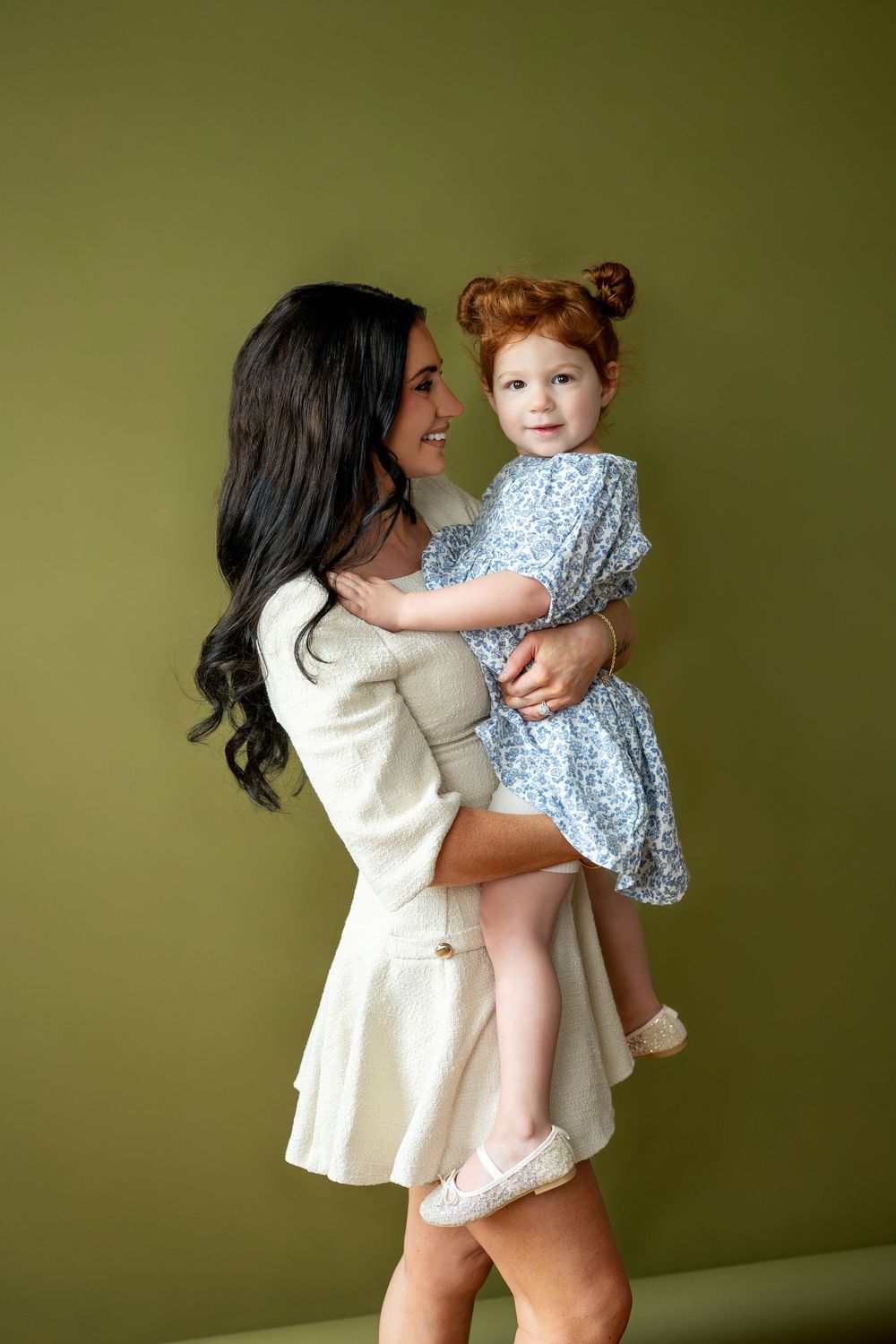 Mother in white dress holding redhead toddler girl in blue floral dress sideways.