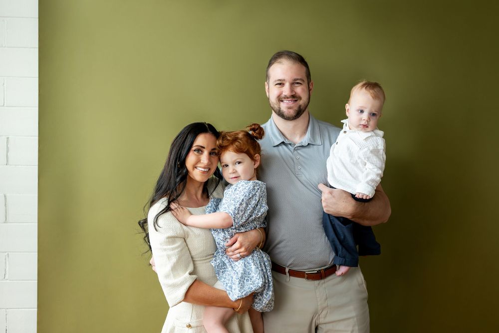 Happy family portrait with brunette mom, redhead toddler, dad, and baby against green wall.