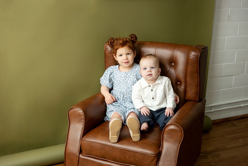Toddler girl with red hair and baby boy in white shirt sitting together on brown leather armchair.