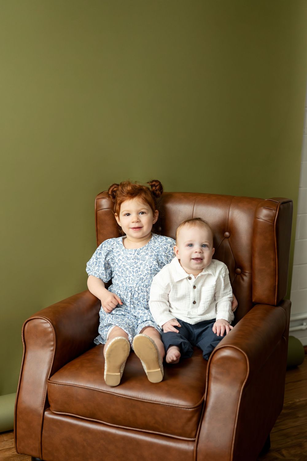 Redhead toddler girl in blue dress and baby boy in white shirt smiling on brown leather chair.